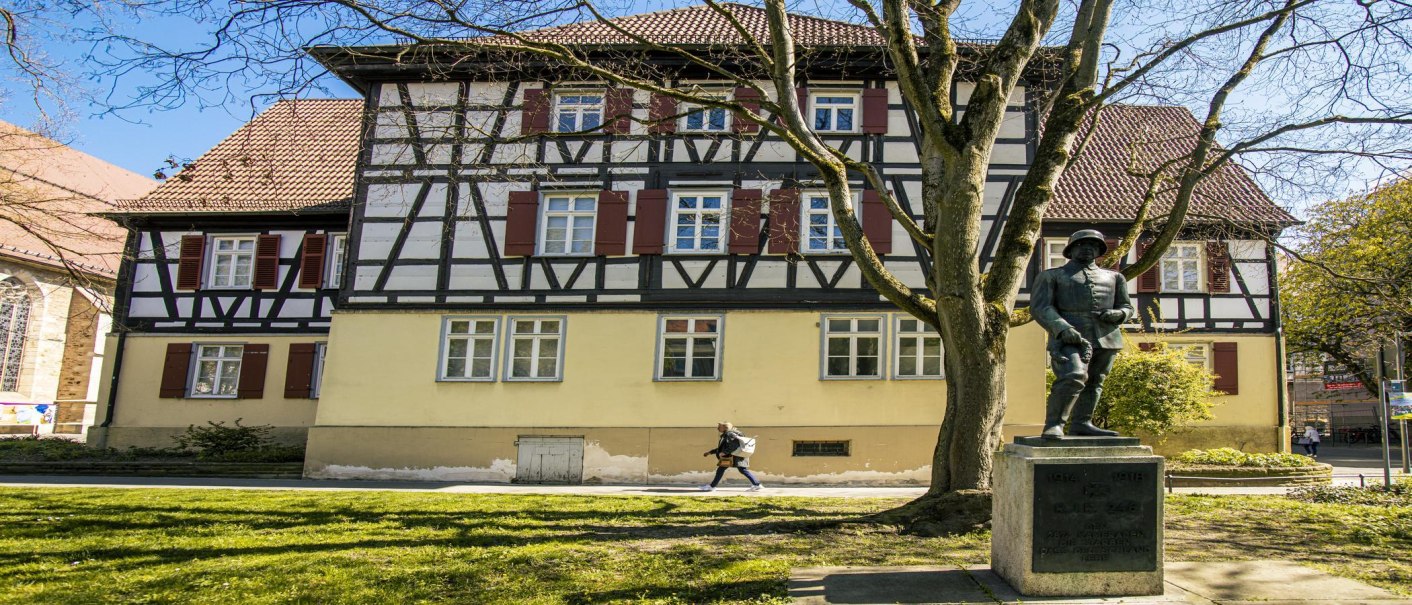 Fachwerkhaus in Kirchheim unter Teck mit einer Statue im Vordergrund. Ein Baum und eine Person, die vorbeigeht, sind ebenfalls zu sehen., © SMG, Sarah Schmid Fachwerkhaus in Kirchheim unter Teck mit einer Statue im Vordergrund. Ein Baum und eine Person, die vorbeigeht, sind ebenfalls zu sehen., © SMG, Sarah Schmid