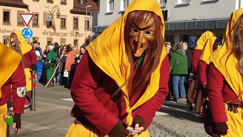 Teilnehmer des Fasnetsumzugs in Kirchheim unter Teck tragen bunte Kostüme und Holzmasken. Zuschauer stehen am Straßenrand vor historischen Gebäuden., © Römer Teilnehmer des Fasnetsumzugs in Kirchheim unter Teck tragen bunte Kostüme und Holzmasken. Zuschauer stehen am Straßenrand vor historischen Gebäuden., © Römer