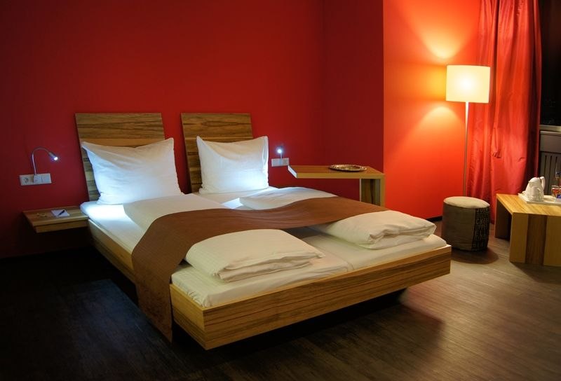 Modernes Hotelzimmer mit roter Wand, Doppelbett, Nachttischlampen und Stehlampe. Elegante Einrichtung mit Holzm&ouml;beln und warmem Licht., &copy; AMH Airport-Messe-Hotel