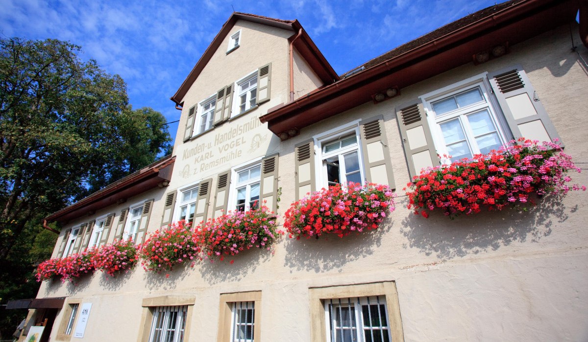 Historisches Gebäude mit weißen Fensterläden und blühenden Blumenkästen vor einem blauen Himmel. Schriftzug "Kunden- u. Handelsmühle Karl Vogel z. Remsmühle" sichtbar., © Remstal Tourismus e.V. Historisches Gebäude mit weißen Fensterläden und blühenden Blumenkästen vor einem blauen Himmel. Schriftzug "Kunden- u. Handelsmühle Karl Vogel z. Remsmühle" sichtbar., © Remstal Tourismus e.V.