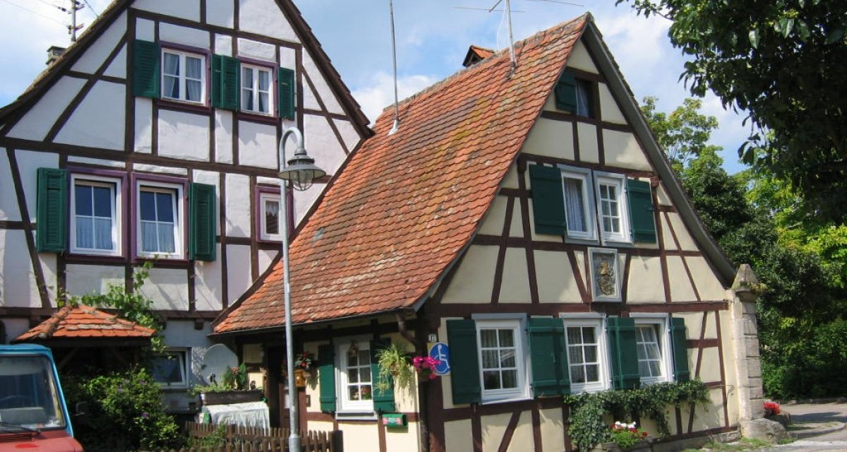 Zwei Fachwerkhäuser mit roten Ziegeldächern und grünen Fensterläden, umgeben von Pflanzen und einem geparkten blauen Fahrzeug., © Land der 1000 Hügel - Kraichgau-Stromberg Zwei Fachwerkhäuser mit roten Ziegeldächern und grünen Fensterläden, umgeben von Pflanzen und einem geparkten blauen Fahrzeug., © Land der 1000 Hügel - Kraichgau-Stromberg