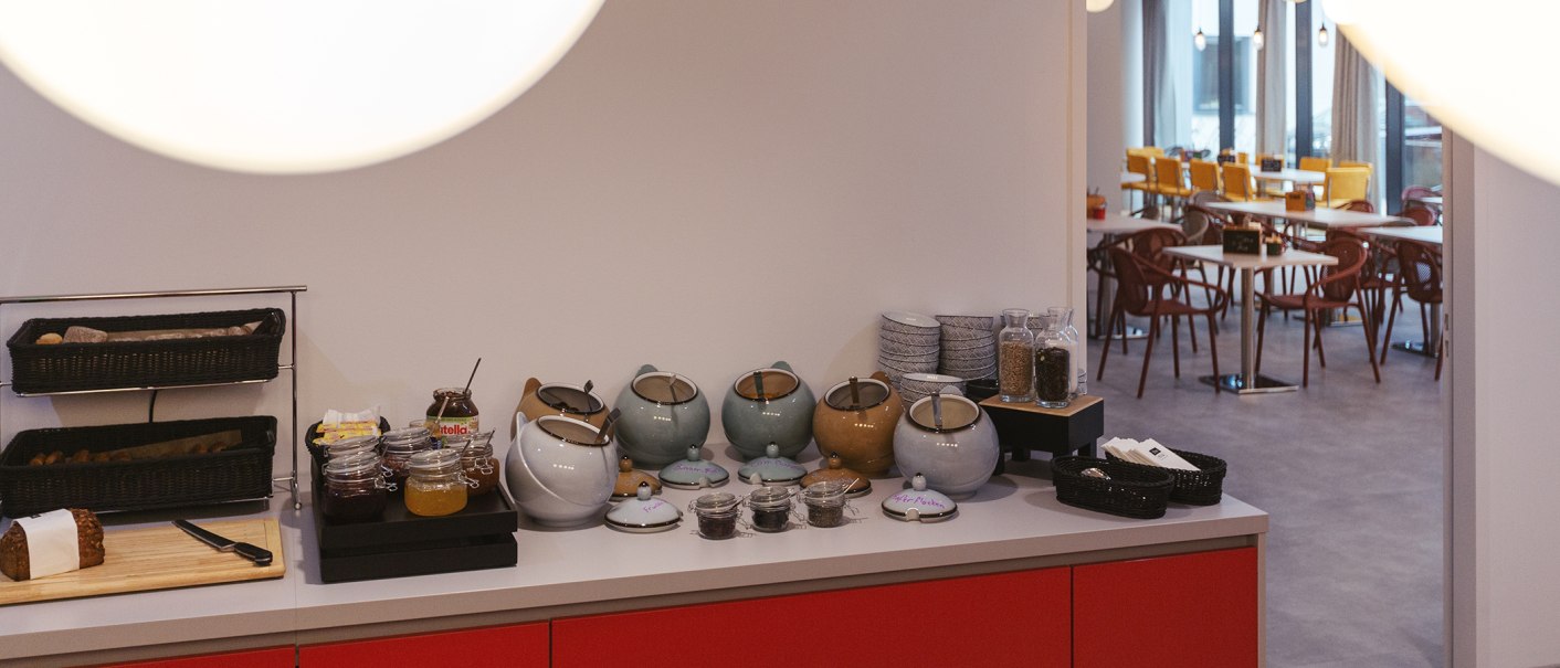 Frühstücksbuffet mit Brotkorb, Marmeladen, Teekannen und Geschirr. Im Hintergrund ein Restaurantbereich mit Tischen und Stühlen., © the niu Form - Novum Hospitality