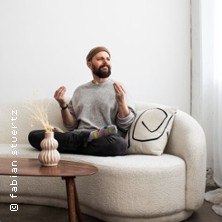 Mann sitzt lächelnd auf einem Sofa, gekreuzte Beine, erhobene Hände. Neben ihm ein Tisch mit Vase. Heller Raum, entspannte Atmosphäre., © links im Bild