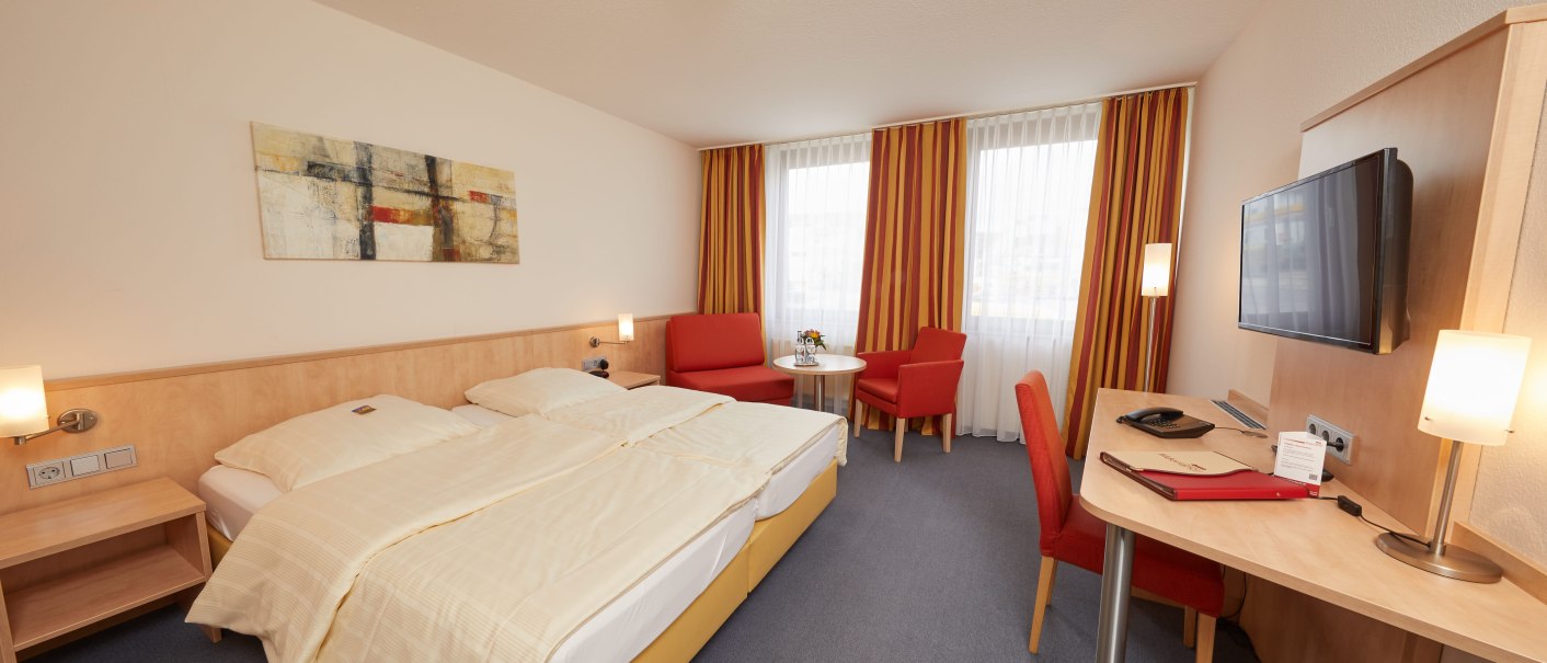 Ein gemütliches Hotelzimmer mit Doppelbett, Schreibtisch, Fernseher und einem kleinen Sitzbereich. Die Vorhänge sind rot-gelb gestreift., © TOMAS