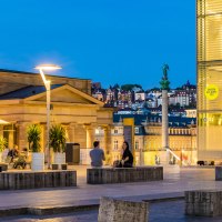 Abendstimmung am Kleinen Schlossplatz mit Palmen, Menschen und moderner Architektur. Im Hintergrund sind beleuchtete Geb&auml;ude und eine Statue zu sehen., &copy; SMG, Werner Dieterich