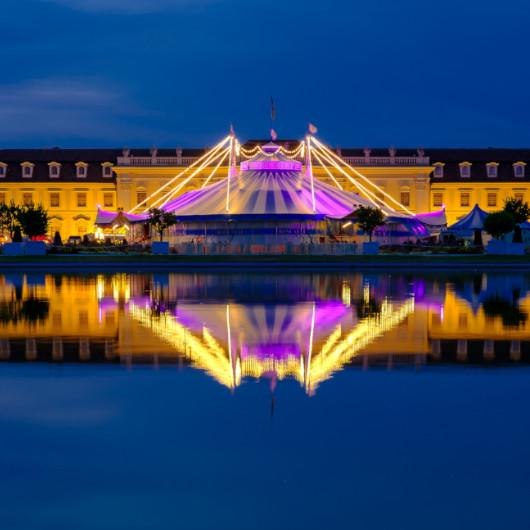 Ein beleuchtetes Zirkuszelt von Circus Roncalli im Bl&uuml;henden Barock, reflektiert in einem Wasserbecken bei Nacht. Die Lichter strahlen in Gelb und Lila., &copy; &copy; Oliver Kelkar