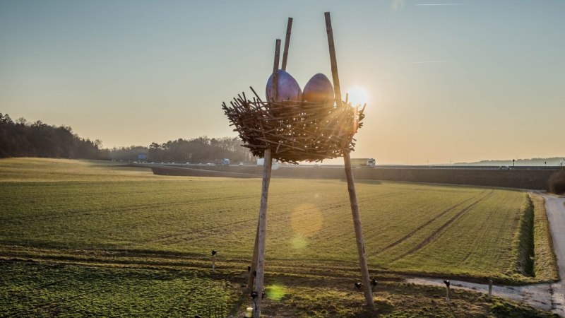 Ein riesiges Vogelnest mit zwei großen Eiern steht auf Stelzen in einer weiten Landschaft. Die Sonne geht im Hintergrund unter., © Natur.Nah. Schönbuch & Heckengäu Ein riesiges Vogelnest mit zwei großen Eiern steht auf Stelzen in einer weiten Landschaft. Die Sonne geht im Hintergrund unter., © Natur.Nah. Schönbuch & Heckengäu