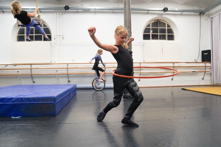 Kinder üben Zirkuskunststücke: Ein Kind mit Hula-Hoop-Reifen, ein anderes auf einem Einrad und ein drittes schwingt an einem Trapez., © Kunstschule Labyrinth