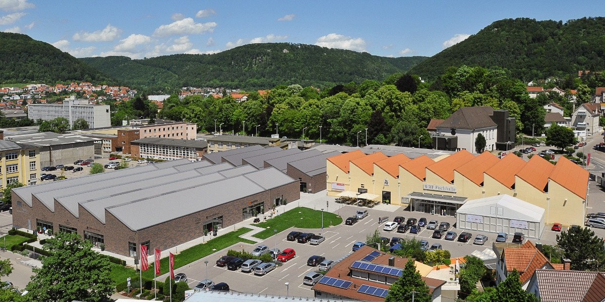 Luftaufnahme des WMF Outlet-Centers mit markanten Dächern, umgeben von grünen Hügeln und einer kleinen Stadt im Hintergrund., © Stuttgart-Marketing GmbH Luftaufnahme des WMF Outlet-Centers mit markanten Dächern, umgeben von grünen Hügeln und einer kleinen Stadt im Hintergrund., © Stuttgart-Marketing GmbH