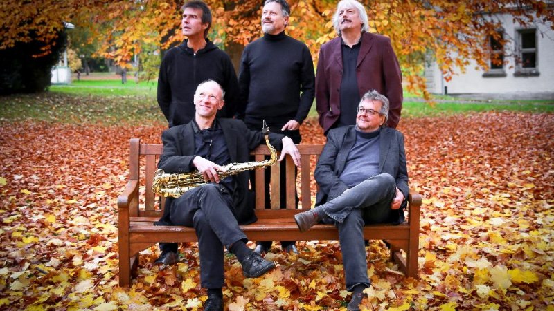 F&uuml;nf M&auml;nner in herbstlicher Umgebung, zwei sitzen auf einer Bank, einer h&auml;lt ein Saxophon. Bunte Bl&auml;tter bedecken den Boden., &copy; Foto Doris Bredow