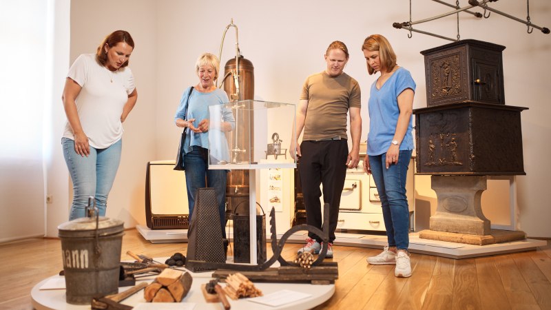 Vier Personen betrachten historische Haushaltsgegenstände im Museum der Alltagskultur. Im Hintergrund sind ein alter Ofen und andere Exponate zu sehen., © FTGRF.de Vier Personen betrachten historische Haushaltsgegenstände im Museum der Alltagskultur. Im Hintergrund sind ein alter Ofen und andere Exponate zu sehen., © FTGRF.de