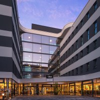 Moderne Hotelaußenfassade mit Glasfront und beleuchtetem Eingangsbereich bei Dämmerung. Das Gebäude hat ein elegantes, wellenförmiges Design., © Novum Hospitality Moderne Hotelaußenfassade mit Glasfront und beleuchtetem Eingangsbereich bei Dämmerung. Das Gebäude hat ein elegantes, wellenförmiges Design., © Novum Hospitality