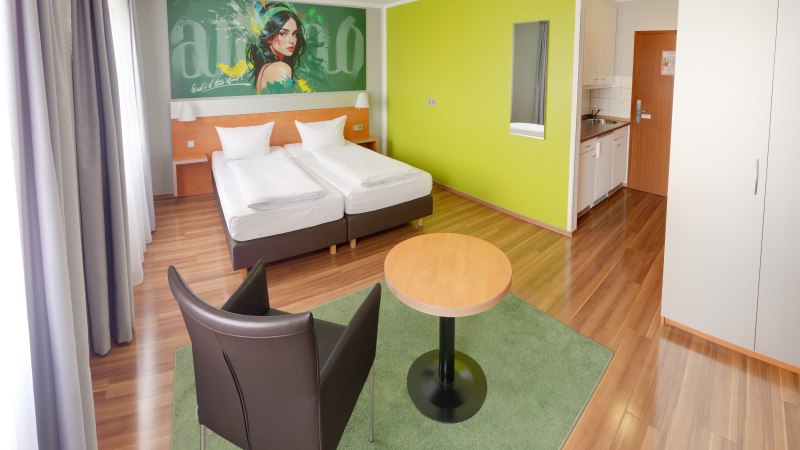 Modernes Superior Doppelzimmer mit zwei Betten, grünem Wandbild, Tisch, Stuhl und kleiner Küchenzeile auf Holzboden., © attimo Hotel Stuttgart