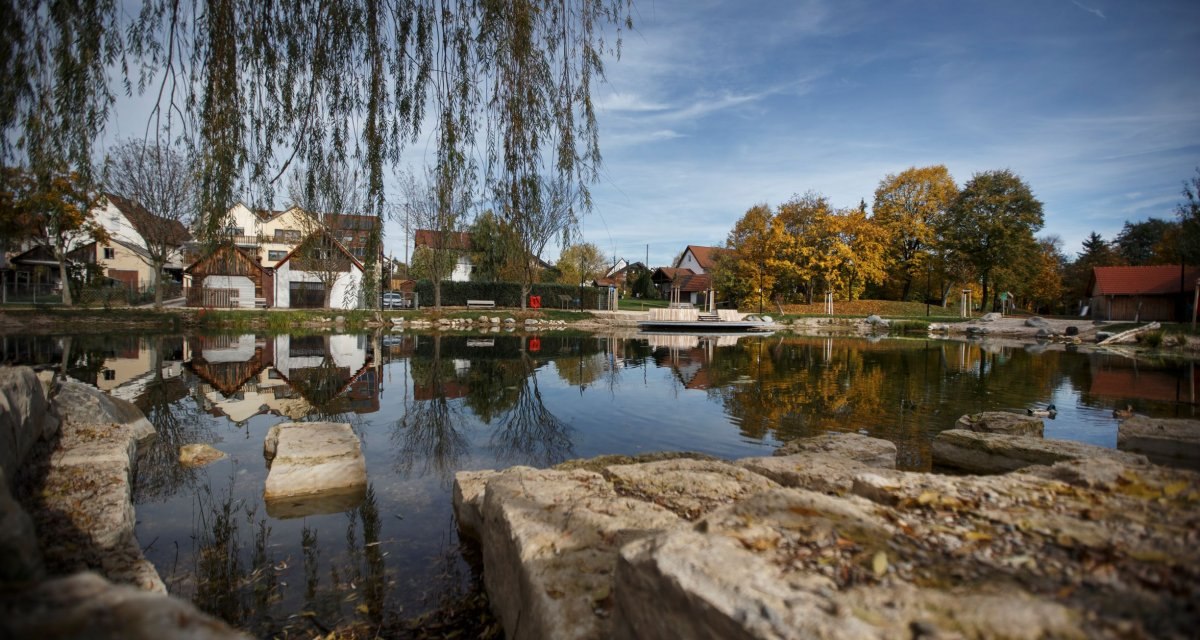 Ein Teich mit Steinen im Vordergrund, umgeben von herbstlichen Bäumen und Häusern. Die ruhige Wasseroberfläche spiegelt die Umgebung wider., © Stoppel Ein Teich mit Steinen im Vordergrund, umgeben von herbstlichen Bäumen und Häusern. Die ruhige Wasseroberfläche spiegelt die Umgebung wider., © Stoppel