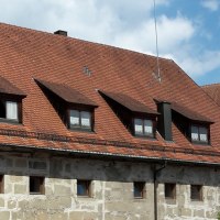 langestrecktes Steingebäude mit kleinen Fenstern und rotbraunem Ziegeldach mit mehreren Dachgauben, im Hintergrund rechts der Fachwerkgiebel des Alten Schlosses mit braunem Holz und weißen Gefachen, © Stadt Gaildorf