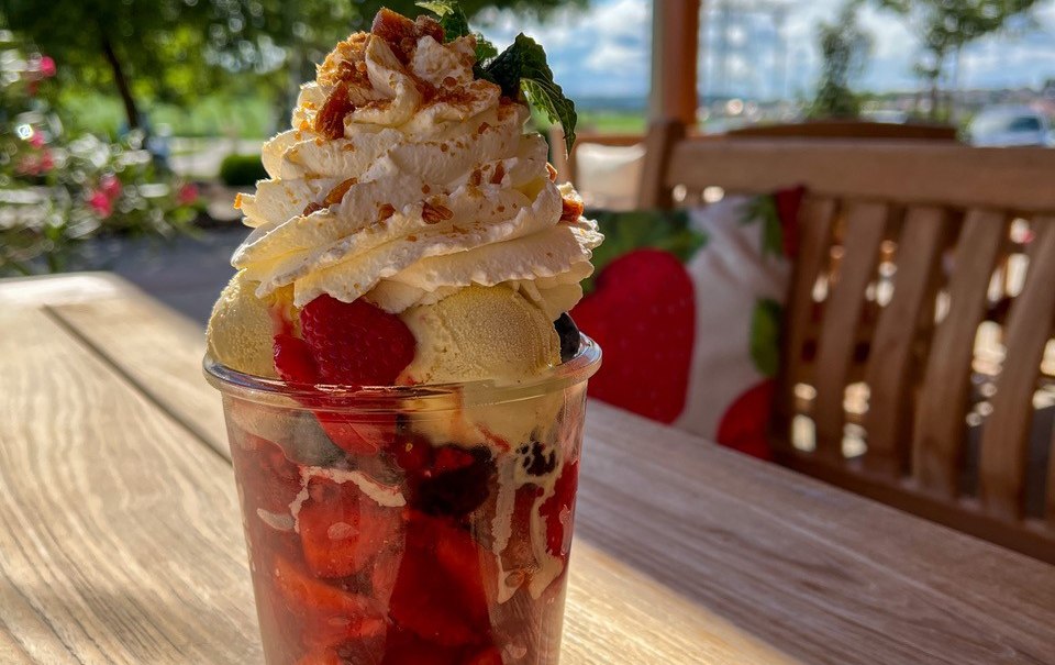 Ein Eisbecher mit Sahne, Erdbeeren und Streuseln steht auf einem Holztisch im Freien. Im Hintergrund sind Bäume und ein blauer Himmel zu sehen., © Engel/ Rammerthof