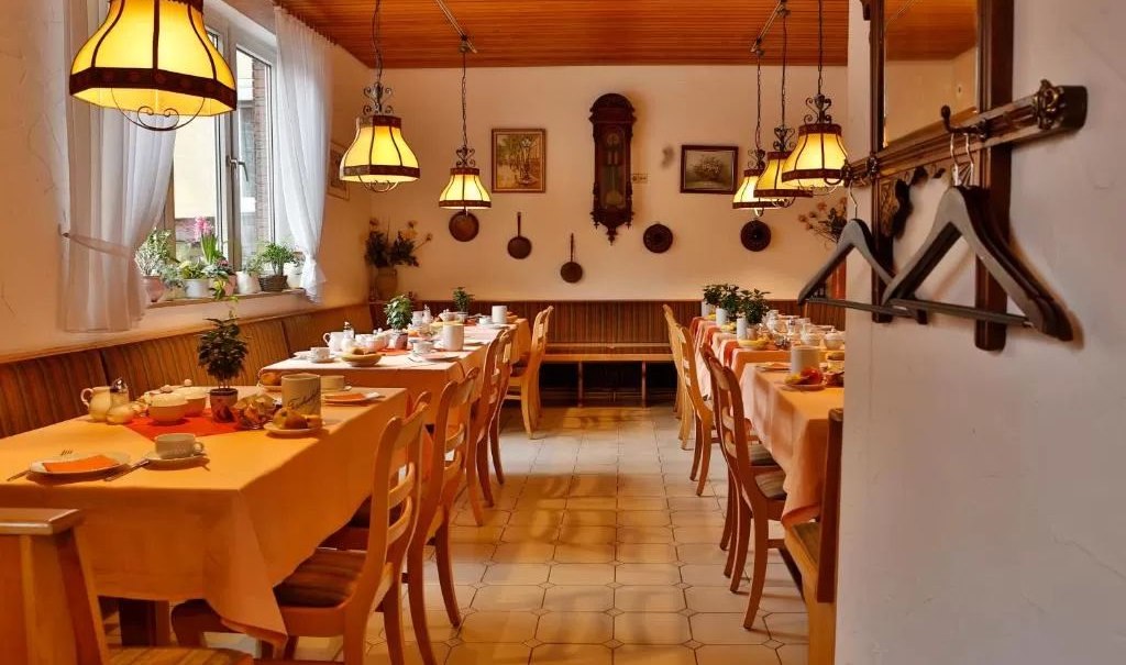 Gemütlicher Frühstücksraum mit langen, gedeckten Tischen, Holzstühlen und warmem Licht von Hängelampen. An der Wand hängen Dekorationen., © Feuerbacher Hof Gemütlicher Frühstücksraum mit langen, gedeckten Tischen, Holzstühlen und warmem Licht von Hängelampen. An der Wand hängen Dekorationen., © Feuerbacher Hof