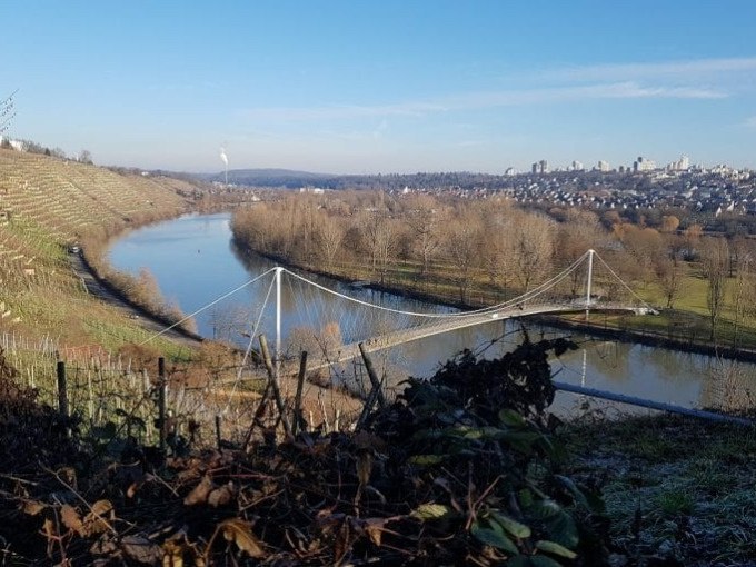 Hängebrücke über einen Fluss, umgeben von Weinbergen und kahlen Bäumen, mit einer Stadt im Hintergrund unter klarem Himmel., © Cool-Tours StattReisen