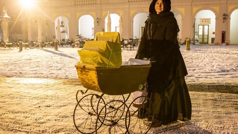 Eine Person in schwarzem Mantel steht mit einem antiken Kinderwagen im Schnee vor einem beleuchteten Gebäude mit Bögen., © Tourist Information und Ticket Service Ludwigsburg Eine Person in schwarzem Mantel steht mit einem antiken Kinderwagen im Schnee vor einem beleuchteten Gebäude mit Bögen., © Tourist Information und Ticket Service Ludwigsburg