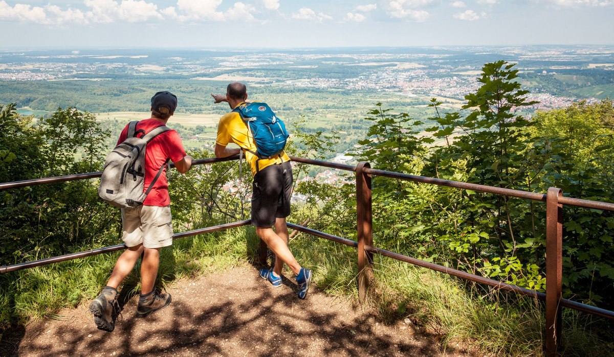 Zwei Wanderer mit Rucksäcken stehen an einem Geländer und blicken auf eine weite, grüne Landschaft unter einem blauen Himmel mit Wolken., © hochgehberge