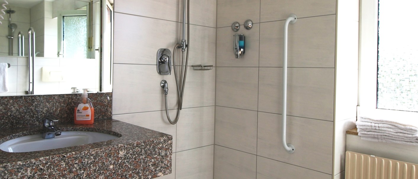 Barrierefreies Badezimmer mit ebenerdiger Dusche, Haltegriff, Waschbecken mit Granitplatte und Handtüchern auf der Fensterbank., © TOMAS Barrierefreies Badezimmer mit ebenerdiger Dusche, Haltegriff, Waschbecken mit Granitplatte und Handtüchern auf der Fensterbank., © TOMAS