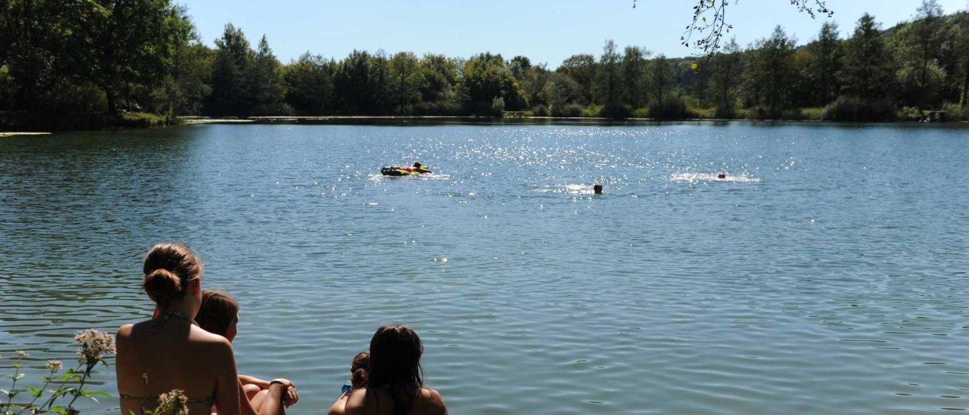 Menschen sitzen am Ufer eines Sees und schauen auf das Wasser, wo andere schwimmen. Die Umgebung ist von B&auml;umen umgeben., &copy; Stadt Vaihingen an der Enz, Foto: Pfisterer