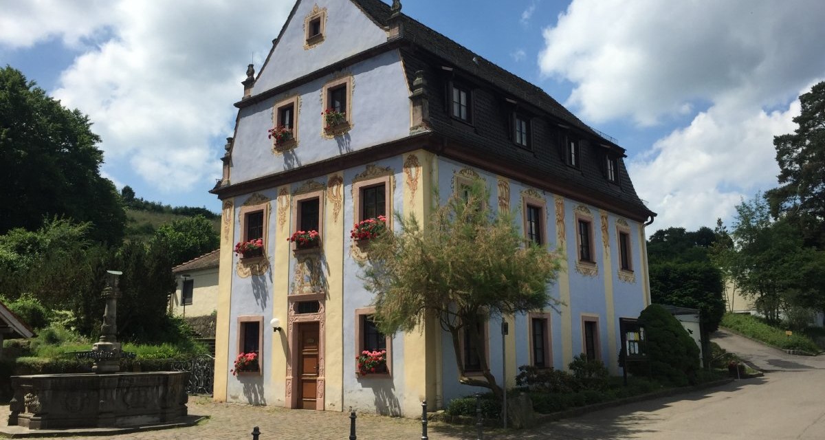 Ein historisches Geb&auml;ude mit verzierten Fenstern und Blumenk&auml;sten, daneben ein Brunnen. Der Himmel ist blau mit einigen Wolken., &copy; www.pro-cycl.de