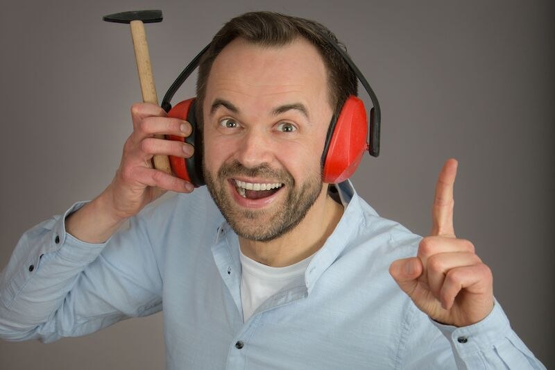 Ein Mann mit roten Ohrenschützern hält einen Hammer ans Ohr und lächelt, während er mit dem Finger nach oben zeigt. Er trägt ein hellblaues Hemd., © Kurverwaltung Bad Urach