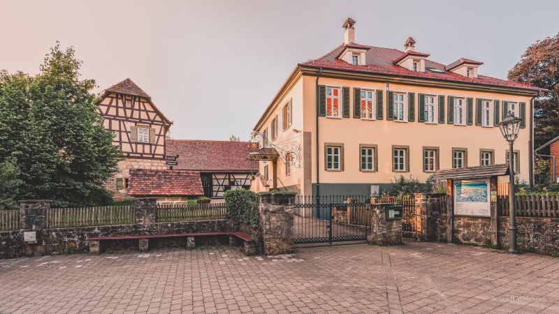 Ein historisches Fachwerkhaus neben einem modernen Gebäude mit grünen Fensterläden in Welzheim. Davor ein gepflasterter Platz und ein schmiedeeisernes Tor., © Stadt Welzheim Ein historisches Fachwerkhaus neben einem modernen Gebäude mit grünen Fensterläden in Welzheim. Davor ein gepflasterter Platz und ein schmiedeeisernes Tor., © Stadt Welzheim
