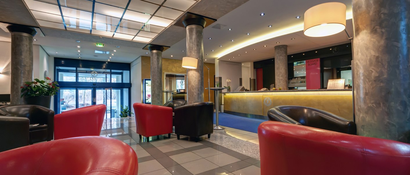 Moderne Lobby mit roten und schwarzen Sesseln, Empfangstresen, Glasdecke und großen Säulen., © City Hotel Reutlingen