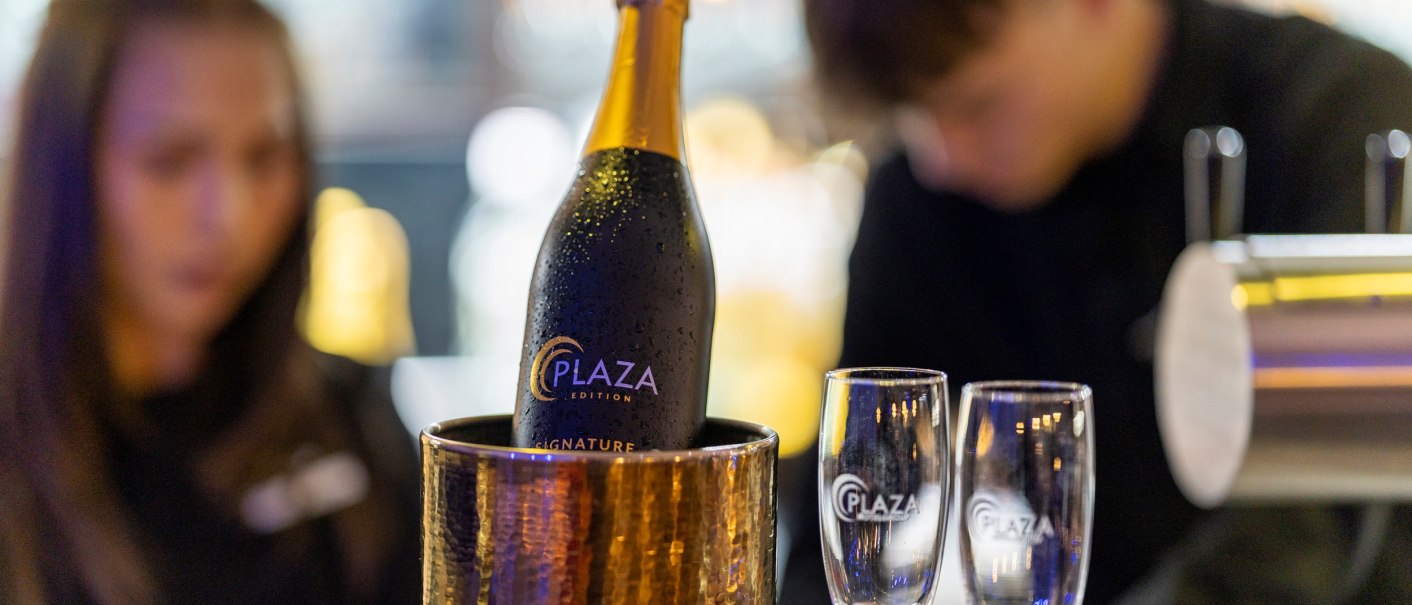 Champagnerflasche 'Plaza Edition' in einem Eiskübel, zwei Gläser daneben. Unscharfe Personen im Hintergrund, Bar-Atmosphäre., © Plaza Residence Stuttgart-Ditzingen Champagnerflasche 'Plaza Edition' in einem Eiskübel, zwei Gläser daneben. Unscharfe Personen im Hintergrund, Bar-Atmosphäre., © Plaza Residence Stuttgart-Ditzingen