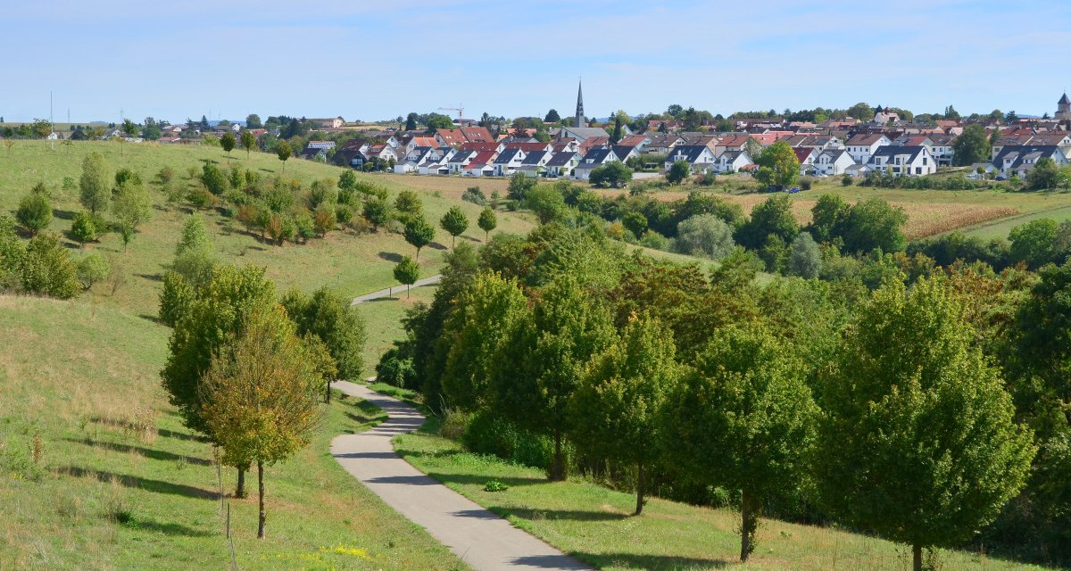 Ein gewundener Weg führt durch eine grüne Landschaft mit Bäumen. Im Hintergrund ist eine Siedlung mit Häusern und einer Kirche sichtbar., © Remstal Tourismus e.V. Ein gewundener Weg führt durch eine grüne Landschaft mit Bäumen. Im Hintergrund ist eine Siedlung mit Häusern und einer Kirche sichtbar., © Remstal Tourismus e.V.