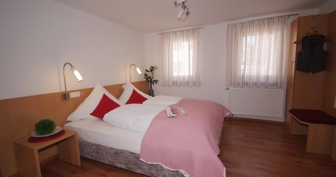 Schlafzimmer mit Doppelbett, rosa Decke, Holzmöbeln und zwei Fenstern mit Vorhängen. Eine Pflanze und ein Magazin auf dem Bett., © TOMAS