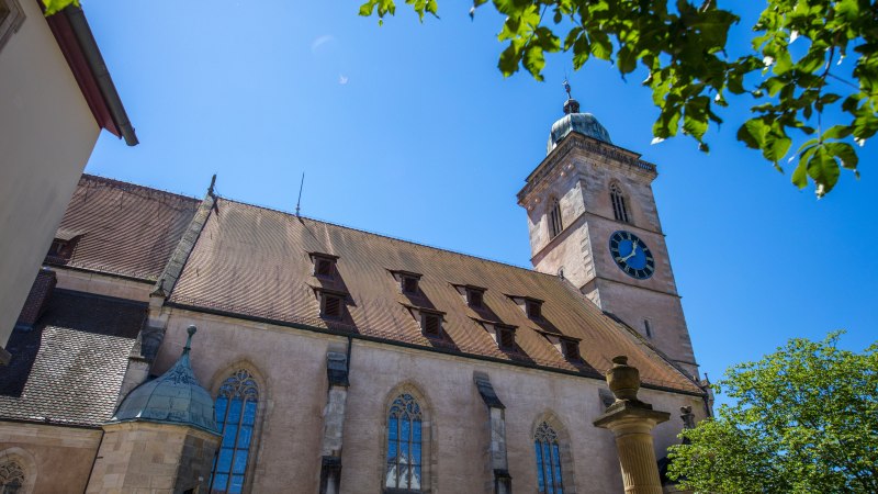 Die Nürtinger Stadtkirche St. Laurentius mit ihrem markanten Turm und einer großen Uhr, umgeben von Bäumen und strahlend blauem Himmel., © SMG, A. Mende