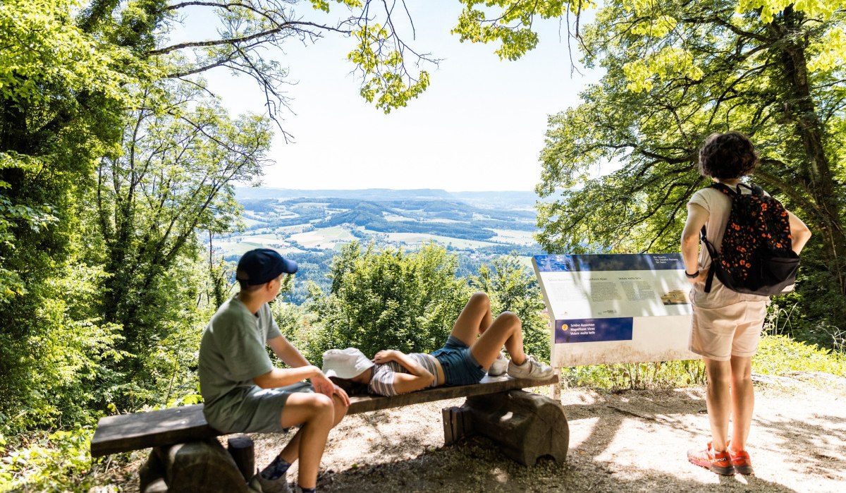 Drei Personen an einem Rastplatz am Hohenstaufen. Zwei sitzen auf einer Bank, eine Person steht und liest eine Infotafel. Im Hintergrund weite Landschaft., © TMBW