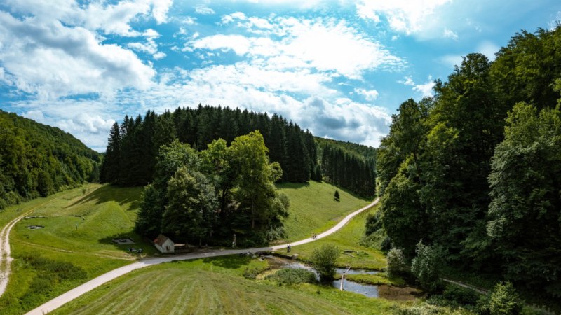 Vogelperspektive auf eine gr&uuml;ne Landschaft mit W&auml;ldern, Wiesen und einem kleinen Bach. Ein Weg schl&auml;ngelt sich durch die Szenerie unter einem blauen Himmel., &copy; Landkreis G&ouml;ppingen