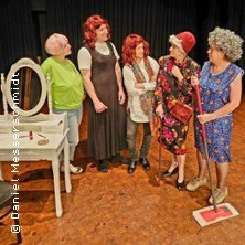 F&uuml;nf Personen in farbenfrohen Kost&uuml;men stehen auf einer B&uuml;hne und unterhalten sich. Eine Person h&auml;lt einen Wischmopp, w&auml;hrend eine andere neben einem Schminktisch steht., &copy; links im Bild