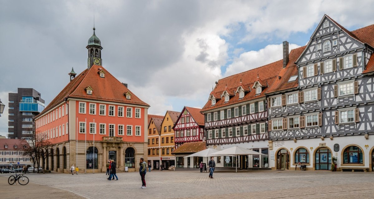Der Marktplatz in Schorndorf zeigt historische Fachwerkh&auml;user und ein rotes Geb&auml;ude mit Turm. Menschen spazieren &uuml;ber den Platz.