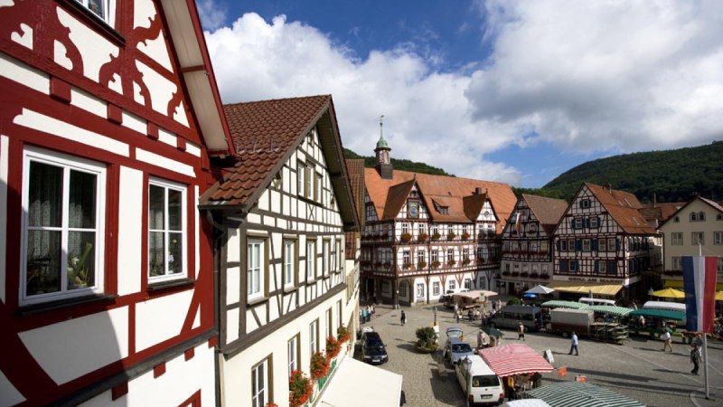Blick auf den Marktplatz in Bad Urach mit malerischen Fachwerkhäusern und bunten Marktständen unter einem blauen Himmel mit Wolken., © Bad Urach Tourismus Blick auf den Marktplatz in Bad Urach mit malerischen Fachwerkhäusern und bunten Marktständen unter einem blauen Himmel mit Wolken., © Bad Urach Tourismus