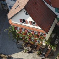 Traditionelles Gebäude mit roten Fensterläden und blumengeschmückter Fassade. Im gepflasterten Hof stehen Tische und Stühle bereit., © cWeinstube Traditionelles Gebäude mit roten Fensterläden und blumengeschmückter Fassade. Im gepflasterten Hof stehen Tische und Stühle bereit., © cWeinstube