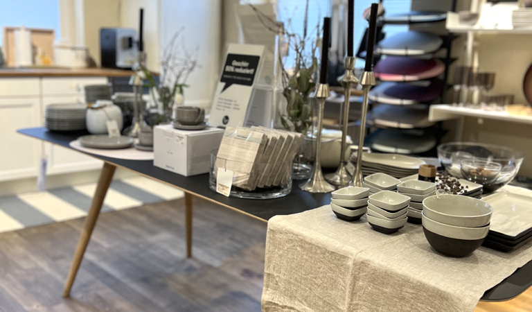 Ein Tisch in einem Laden mit Geschirr, Schalen, Kerzenhaltern und Vasen. Der Raum hat Holzboden und weiße Wände mit Regalen., © schöner laden