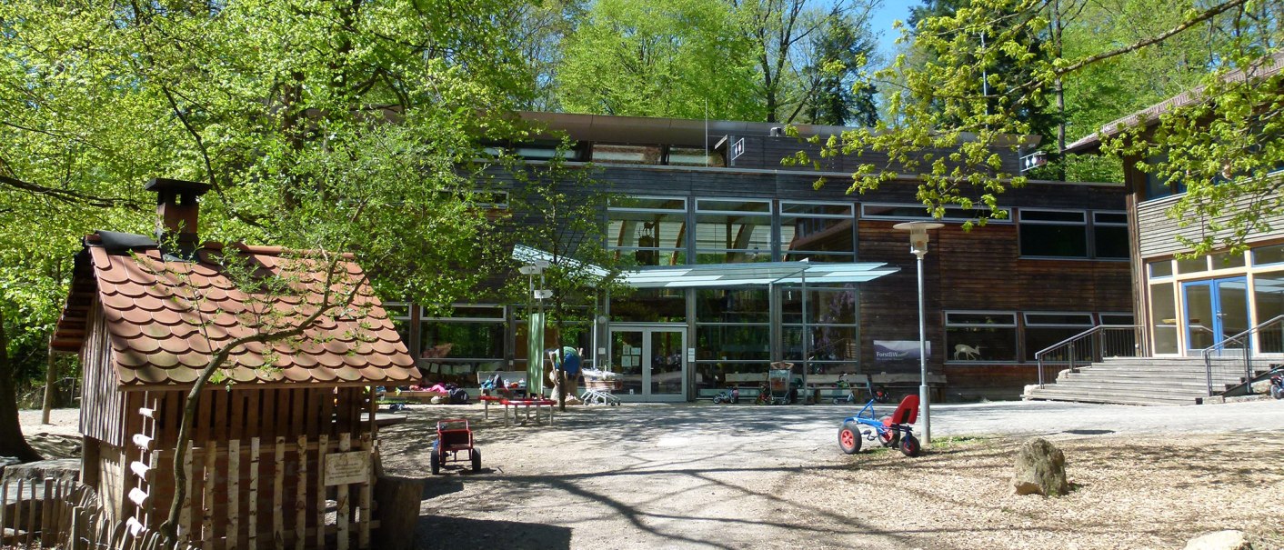 Innenhof des Haus des Waldes mit Spielzeugfahrzeugen, Bäumen und einem kleinen Holzhaus. Moderne Glasfassade im Hintergrund., © Haus des Waldes