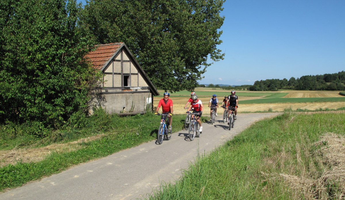 Vier Radfahrer fahren auf einem ländlichen Weg an einem kleinen Fachwerkhaus vorbei, umgeben von Feldern und Bäumen unter blauem Himmel., © Natur.Nah. Schönbuch & Heckengäu Vier Radfahrer fahren auf einem ländlichen Weg an einem kleinen Fachwerkhaus vorbei, umgeben von Feldern und Bäumen unter blauem Himmel., © Natur.Nah. Schönbuch & Heckengäu
