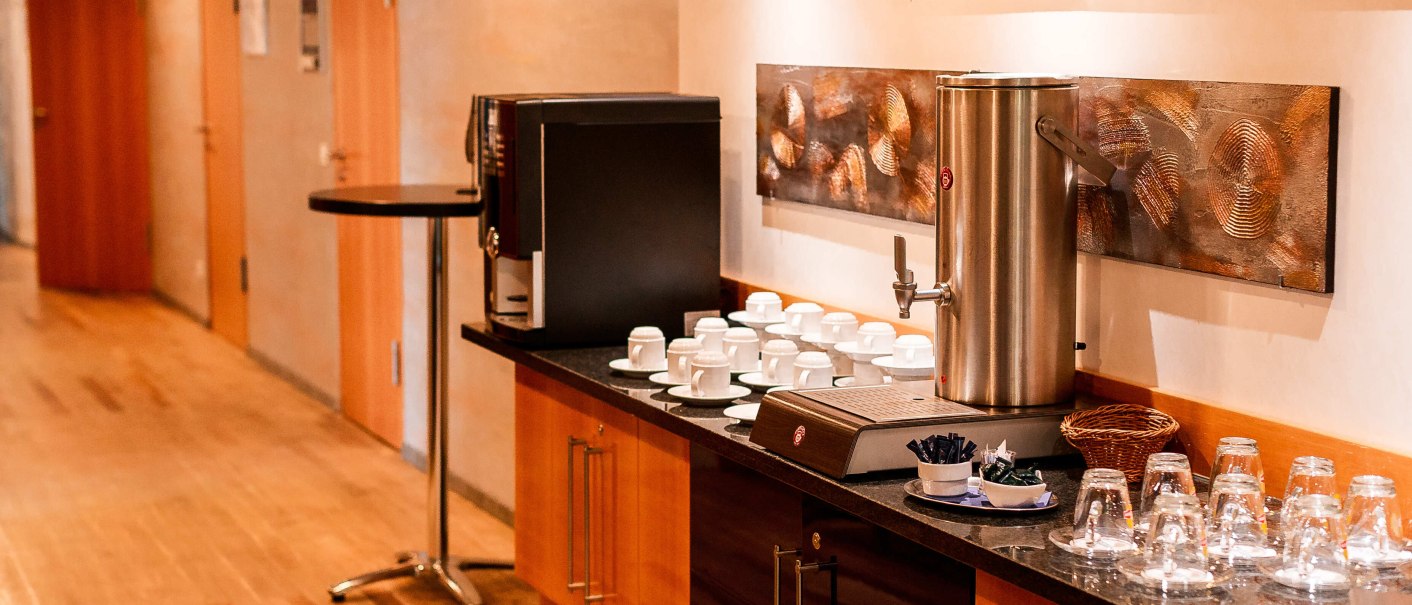 Kaffeestation in einem Flur mit Tassen, Gläsern und Kaffeemaschine auf einem Holztresen. Kunstwerke an der Wand., © Plaza Residence Stuttgart-Ditzingen Kaffeestation in einem Flur mit Tassen, Gläsern und Kaffeemaschine auf einem Holztresen. Kunstwerke an der Wand., © Plaza Residence Stuttgart-Ditzingen