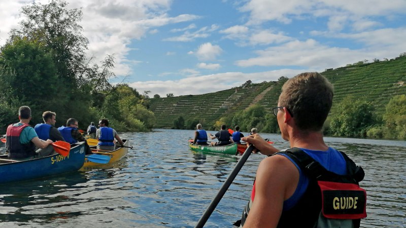 Eine Gruppe von Menschen paddelt in Kanus auf einem Fluss, umgeben von grünen Hügeln. Ein Guide ist im Vordergrund zu sehen., © Die Zugvögel