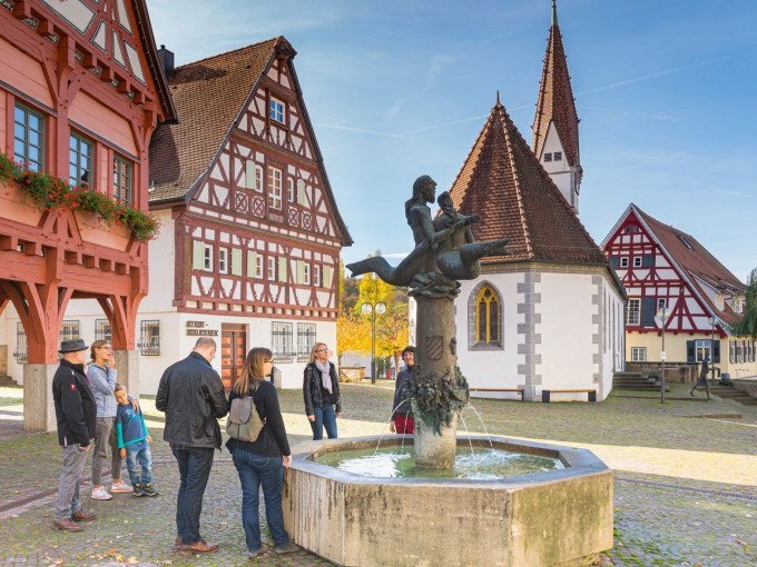 Eine Gruppe von Menschen betrachtet einen Brunnen auf einem historischen Platz in Plochingen. Fachwerkh&auml;user und eine Kirche im Hintergrund., &copy; PlochingenInfo