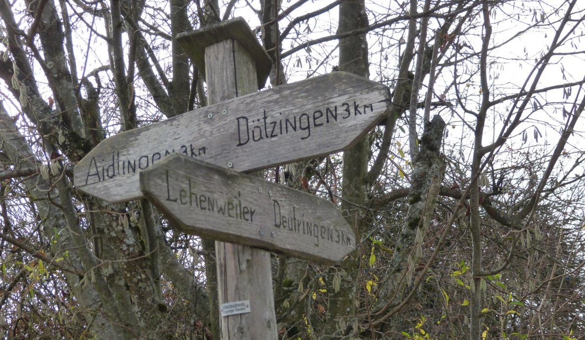 Ein h&ouml;lzerner Wegweiser im Wald zeigt Richtungen zu D&auml;tzingen, Aidlingen und Lehenweiler an. Ein Schild mit gelber Raute ist darunter angebracht., &copy; Natur.Nah. Sch&ouml;nbuch & Heckeng&auml;u