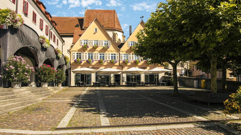 Marktplatz in Bietigheim-Bissingen mit historischen Gebäuden, Blumen und Sonnenschirmen vor einem Café. Ein Baum spendet Schatten., © Stuttgart Marketing GmbH, Sarah Schmid