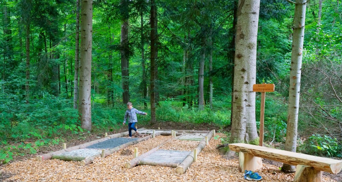 Ein Kind läuft auf einem Barfußpfad im Wald. Der Pfad besteht aus verschiedenen Materialien. Eine Holzbank und ein Schild sind sichtbar., © Touristikgemeinschaft Hohenlohe e. V. | Stadt Künzelsau Ein Kind läuft auf einem Barfußpfad im Wald. Der Pfad besteht aus verschiedenen Materialien. Eine Holzbank und ein Schild sind sichtbar., © Touristikgemeinschaft Hohenlohe e. V. | Stadt Künzelsau