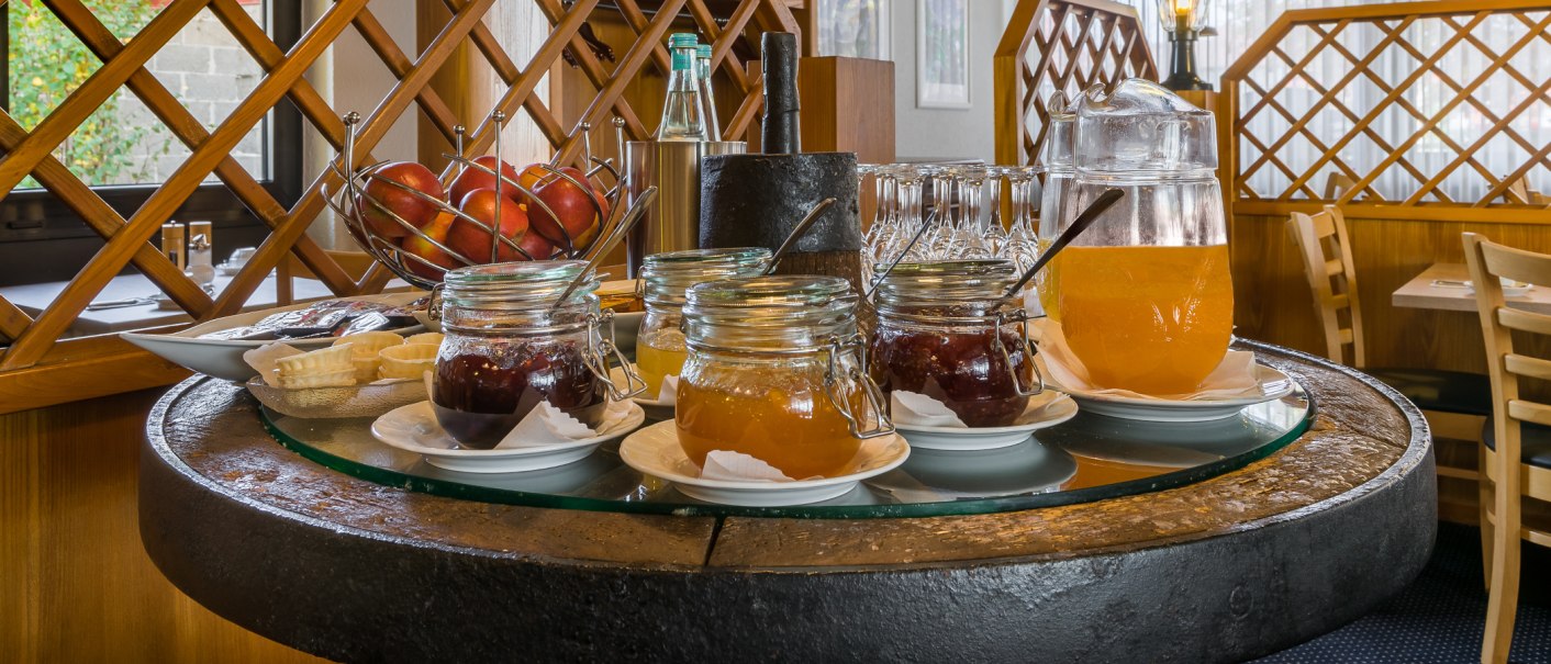 Ein Frühstückstisch mit Marmeladengläsern, einem Krug mit Orangensaft und einem Obstkorb voller Äpfel, umgeben von Holzmöbeln., © Novum Management GmbH